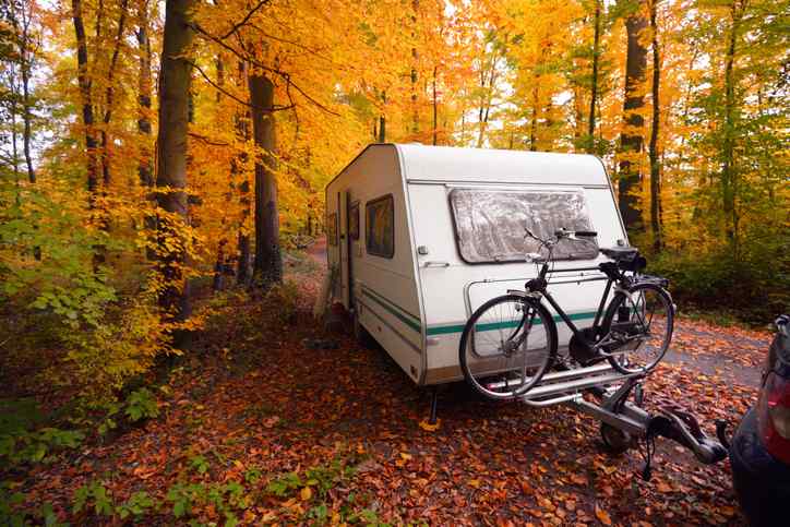 Caravan trailer with a bicycle and a car parked among autumn yellow trees.