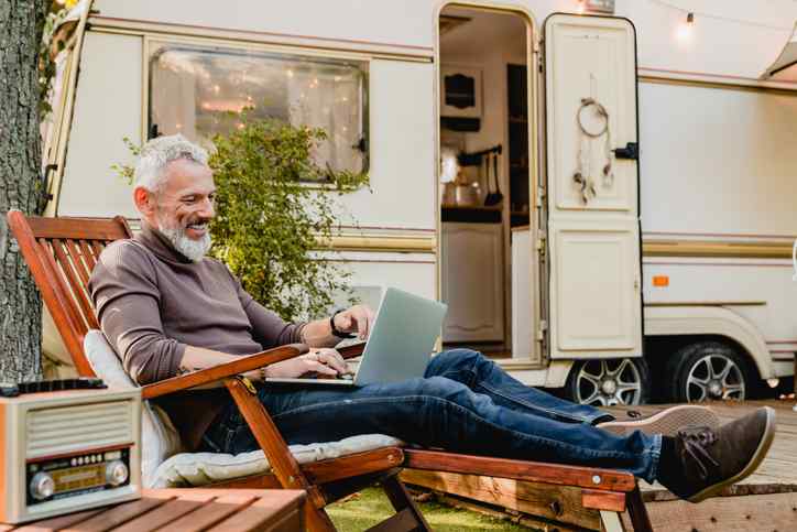 Relaxed grey-haired man resting on the wooden deck chair using laptop with camping van in background.